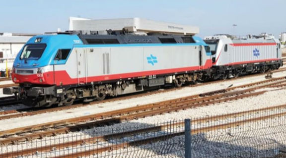Locomotora con colores gris, azul y rojo, unida al primer vagón, en segunda fila de vías de tren.